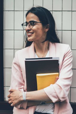 Ellerinde eğitim teçhizatıyla mutlu, zeki bir bayan öğrenci şehir ortamına bakıyor ve gülümsüyor, gözlüklü muhteşem bir kadın dışarıda gülüyor.