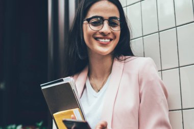 Modern cep telefonu cihazına sahip neşeli nüfuz blogcusu ve içerik yayını için ders kitabı harcamak için boş vakti olan, modaya uygun gözlüklü samimi bir kadın.