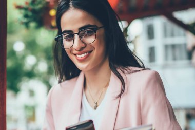 Modern cep telefonu cihazına sahip neşeli nüfuz blogcusu ve içerik yayını için ders kitabı harcamak için boş vakti olan, modaya uygun gözlüklü samimi bir kadın.