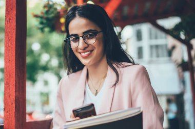 Modern cep telefonu cihazına sahip neşeli nüfuz blogcusu ve içerik yayını için ders kitabı harcamak için boş vakti olan, modaya uygun gözlüklü samimi bir kadın.