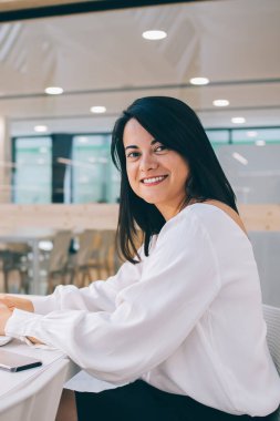 Modern ofiste sandalyede oturan ve kameraya gülümserken boş ekranla dizüstü bilgisayarda çalışan beyaz bluzlu mutlu genç bayan.