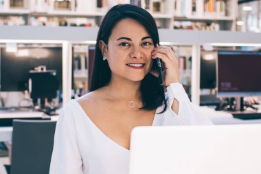 Resmi olarak giyinmiş, siyah saçlı bir kadın çalışan gülümseyip kameraya bakarken cep telefonundan arıyor ve açık ofiste dizüstü bilgisayar kullanıyor.