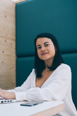 Barcelona 'da modern bir kafede dizüstü bilgisayarda gezinirken gülümseyen ve kameraya bakan beyaz bluzlu muhteşem siyah saçlı serbest kadın.