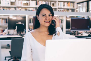 Resmi olarak giyinmiş, siyah saçlı bir kadın çalışan gülümseyip kameraya bakarken cep telefonundan arıyor ve açık ofiste dizüstü bilgisayar kullanıyor.