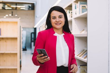 Zarif ceketli genç bir bayan kitaplığın önünde parlak bir kütüphanede duruyor ve kameraya gülümseyerek bakarken cep telefonu kullanıyor.