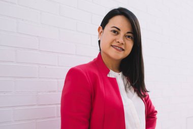 Resmi giyinmiş, renkli pembe ceketli, zarif siyah saçlı kadın Barcelona 'daki beyaz duvarın önünde gülümseyip kameraya bakıyor.