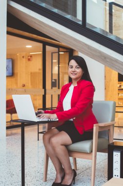 Modern ofiste çalışırken ve kameraya bakarken resmi giyinmiş siyah saçlı, etnik bir kadın masada oturuyor ve netbook 'ta sörf yapıyor.