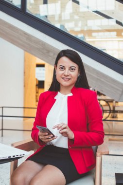 Genç Latin kökenli, siyah saçlı, cep telefonuyla yazı yazan, modern ofiste çalışırken masasında beyaz dizüstü bilgisayarıyla oturan bir kadın.