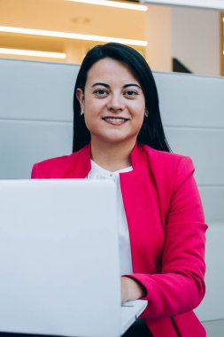 Resmi giysiler içinde genç İspanyol bayan kanepeye oturmuş, bilgisayar başında, modern ofiste proje üzerinde çalışıyor ve kameraya bakıyor.