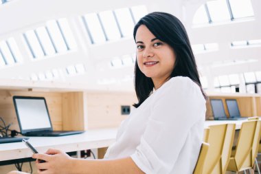 Resmi giysiler içinde gülümseyen bir kadının yan görüntüsü. Akıllı telefonuyla mesajlaşıyor ve kameraya bakarken sandalyede oturuyor.
