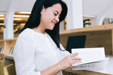 Modern kütüphanede güneşli bir günde masa başında otururken keyifli bir şekilde kitap okuyan neşeli genç bayanın yan görüntüsü.