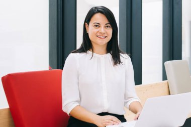 Genç bayan girişimci resmi kıyafetler içinde modern iş yerinde beyaz netbook 'un önündeki pencerenin yanındaki tahta bankta oturuyor.