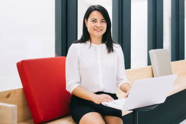 Genç bayan girişimci resmi kıyafetler içinde modern iş yerinde beyaz netbook 'un önündeki pencerenin yanındaki tahta bankta oturuyor.