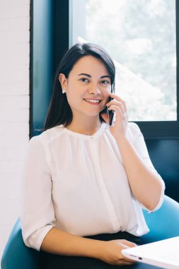 Zarif kıyafetler giymiş, masada dizüstü bilgisayarla oturup kameraya bakarken telefonla iletişim kuran genç gülümseyen yönetici asistanı.