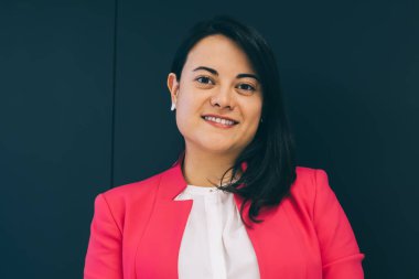 Zarif bir ceket giyen, akıllı telefondan gülümseyen ve internette sörf yapan bayan patron ofis mavi duvarının arkasında kameraya bakıyor.