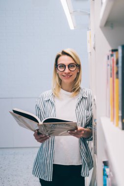 Barcelona 'da üniversite kütüphanesinde kitaplığın yanında durup kitap okurken gülümseyen ve kameraya bakan gözlüklü mutlu genç öğrenci