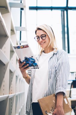 Mağazanın raflarının yanında ilginç açık kitaplarla duran mutlu beyaz kadın ve gülümseyen, eğitim edebiyatı okuyan zeki, sıradan bir kadın kütüphanede ödev için ders kitapları seçiyor.