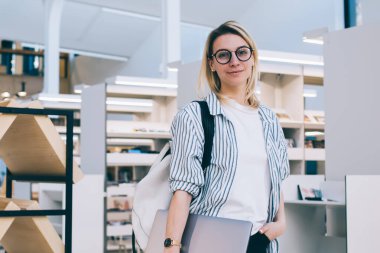 Kitabevinde dizüstü bilgisayarı olan başarılı beyaz kadın ressamın yarım boy portresi, 20 yaşında genç bir öğrenci çalışma alanındaki kitaplığın yanındaki kameraya bakıyor.