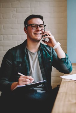 Neşeli erkek serbest meslek sahibi iyi ruh hali planlayıcısı yazma eğitimi sırasında smartphone konuşmayı kafe iç, mutlu hipster adam çalışan proje hakkında telefon paylaşım iyi haber verme