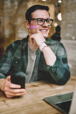 Neşeli hipster adam uzakta smartphone, müreffeh erkek freelancer mesafesi iş modern aygıtlar ve kablosuz internet ile kafeterya iç memnun tutan penceresinde arıyorum gözlükler