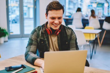 Neşeli hipster adamla erkek öğrenci e öğrenme ders çalışmak için tarama netbook ile zevk gülümseyen eğlence alanı, coworking içinde dizüstü bilgisayar ile harcama bilgisayar oyunları oynarken zevk kulaklık