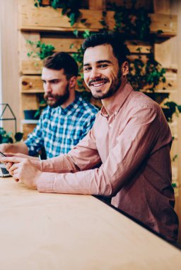 Kameraya bakan neşeli hipster adamın portresi iş arkadaşı ile birlikte çalışma alanında dizüstü bilgisayar kullanarak zengin genç girişimciler birlikte serbest çalışarak para kazanıyorlar.