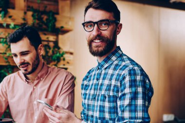 Neşeli bir erkek, kameraya bakıyor ve telefonu tutuyor. Arkadaşı da bilgisayardan internetten bilgi okuyor. Refah içindeki bir çalışan, projedeki meslektaşıyla iş birliği yapıyor.