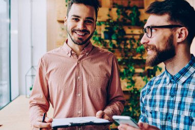 Klasör başarılı başlangıç eğitim hakkında arkadaşıyla danışmanlık sırasında kameraya bakarak hipster adam gülümseyen meslektaşı ile işbirliği için planları ile tutarak yakışıklı neşeli adam portresi