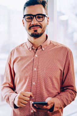 Sıradan giyinen, akıllı telefon takan, kameraya bakan, sosyal ağlarda sohbet etmek için telefonu kullanan, gözlüklü, yakışıklı hippi bir genç girişimcinin yarım boy portresi.