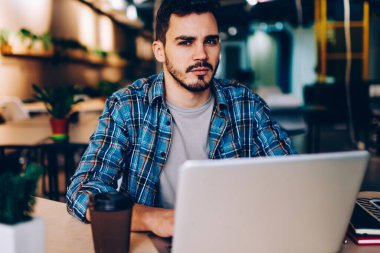 Üniversitede internet ve kablosuz internet kullanarak sınav hazırlığı yapan ciddi bir hipster çocuğun portresi. Kameraya bakan genç adam, birlikte çalıştığı yerdeki dizüstü bilgisayarları kullanarak hayal kırıklığına uğradı.