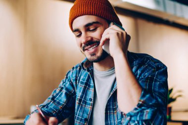 Neşeli erkek yaratıcı tasarımcı defterine çizim yapıyor telefon konuşması yapıyor, gülümseyen hipster adam üniversite kütüphanesinde otururken fikir alışverişi yaptığı için arkadaşına telefon ediyor.