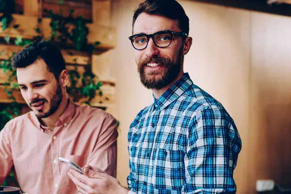 Neşeli bir erkek, kameraya bakıyor ve telefonu tutuyor. Arkadaşı da bilgisayardan internetten bilgi okuyor. Refah içindeki bir çalışan, projedeki meslektaşıyla iş birliği yapıyor.