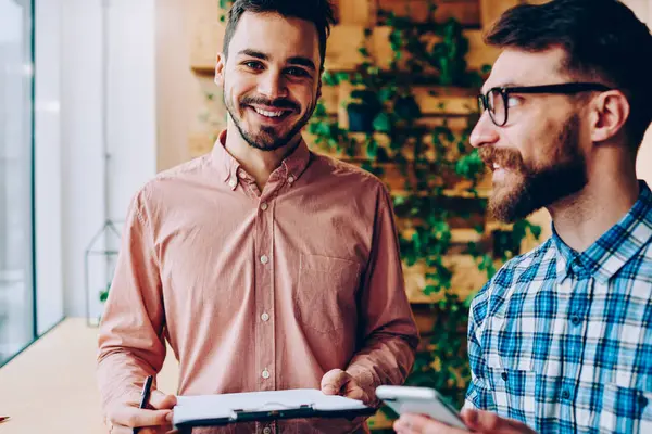 Klasör başarılı başlangıç eğitim hakkında arkadaşıyla danışmanlık sırasında kameraya bakarak hipster adam gülümseyen meslektaşı ile işbirliği için planları ile tutarak yakışıklı neşeli adam portresi