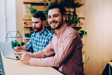 Kameraya bakan neşeli hipster adamın portresi iş arkadaşı ile birlikte çalışma alanında dizüstü bilgisayar kullanarak zengin genç girişimciler birlikte serbest çalışarak para kazanıyorlar.