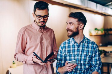 Neşeli hipster adam meslektaşı gülümseyen iletişim iş smartphone, bilgi iş arkadaşınız ile işbirliği için tartışmaya ile adam holding Not defteri kullanarak planlama hakkında ofiste bakarak
