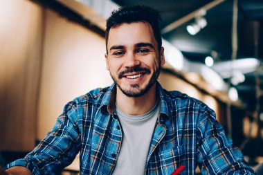 Birlikte çalışmayı seven neşeli erkek öğrencinin portresi araştırma için dizüstü bilgisayarı kullanan mutlu bir serbest yazar, uzaktan yapılan bir iş için fotoğraf çekerken kameraya bakıyor ve notlar alıyor.