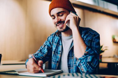 Neşeli erkek yaratıcı tasarımcı defterine çizim yapıyor telefon konuşması yapıyor, gülümseyen hipster adam üniversite kütüphanesinde otururken fikir alışverişi yaptığı için arkadaşına telefon ediyor.