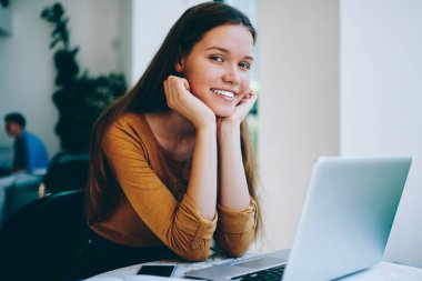 Üniversite kafeteryasında bilgisayar başında çalışırken yüzünde sevimli bir gülümsemeyle kameraya bakan başarılı kadın serbest yazar portresi. Kapalı alanda teknoloji kullanarak okuyan mutlu hippi kız.