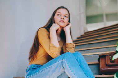 Kot pantolon giymiş, düşünceli bir hippi kız üniversite merdivenlerinde eski moda sırt çantasının yanında oturmuş, şık giyinmiş, boş zamanın tadını çıkarıyor.