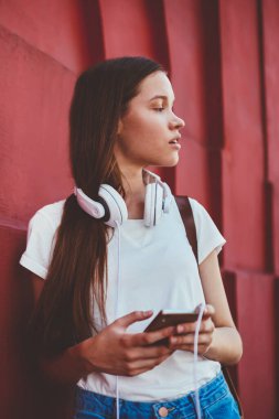 Yoğunlaşmış kadın meloman, yeni cep telefonuyla aramasını beklerken düşünceli bakışlar atıyor. Kafkasyalı kadın, modern akıllı telefonuyla duvarın yanında duruyor. Müzik uygulaması ve kulaklık için.