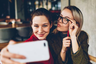 Casual giyim poz ve selfie fotoğraflar yayın için modern smartphone cep telefonu ile sosyal ağlar kafede dostu toplantı sırasında iç yapım komik suratlar ile iki başarılı kadın blogcular