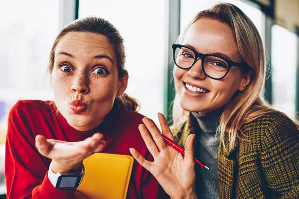 Yarım uzunluk portre pozitif öğrencinin hava öpücük ve güzel sarışın hipster blogger birlikte rekreasyon vaktinizi el sallayarak gözlük gönderme. Mutlu genç kadın eğlenceli kameraya gülümseyen
