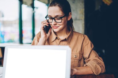 Esmer kadın Yöneticisi smartphone üzerinde arama gözlük '. Freelancer işleci ile mobil faydaları sırasında telefonla söz Eyewear dizüstü bilgisayarda boş bir ekran ile uzaktan çalışma