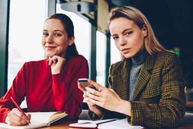 Yarım uzunluk portre iki başarılı tasarımcıların fikirler Smartphone cep telefonu ile arama ve kroki teamworking coworking içinde çizim yaparken kameraya bakıyor. Mobil ve olumlu öğrenci ile sarışın öğretmen