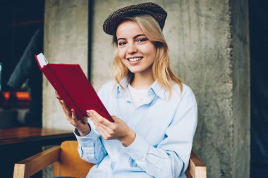 Ders kitabı ile yaklaşan sınavlar için hazırlarken kameraya gülümseyen şık şapka neşeli sarışın yetenekli öğrencinin yarım uzunluk portre. En sevdiğim bestseller zevk başarılı hipster öğrenci