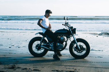 Güneş gözlüklü şık bir adam boş kumlu sahilde park etmiş motosikletli mavi gökyüzünde dalgalı bir sahil kenarında.