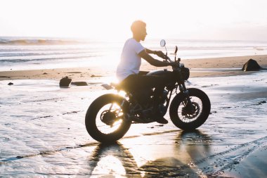 Plaj boyunca motosiklet süren ve deniz manzaralı açık havada gün batımında ıslak kumda lastik izleri bırakan yüzü olmayan bir adamın yan görüntüsü.