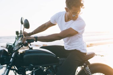 İspanyol yakışıklı, gözlüklü, açık renk elbiseli motosikletli deniz kıyısındaki çölde motosiklet süren ve aşağıya bakan motosikletli bir erkek.