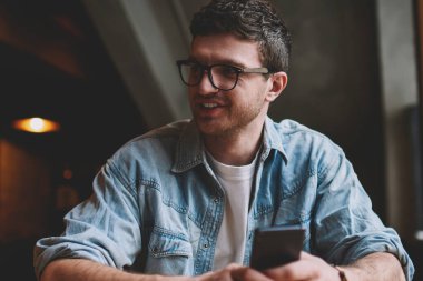 Sohbet etmek için uygulama kurarken elinde cep telefonu tutan sevimli gülümsemeli yakışıklı hippi adam. Gözlüklü neşeli erkek blogcu başka yere bakıyor ve akıllı telefon aygıtı kullanıyor.