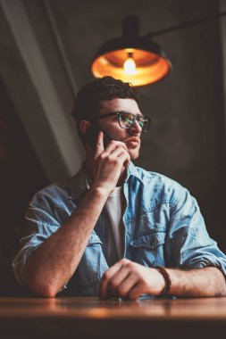 Genç konsantre hipster adam önemli bir akıllı telefon konuşması sırasında düşünceli bir şekilde yana bakıyor, ciddi bir beyaz adam arkadaşını arıyor mobil uygulama ve kapalı alandaki kablosuz bağlantıyı kullanıyor.
